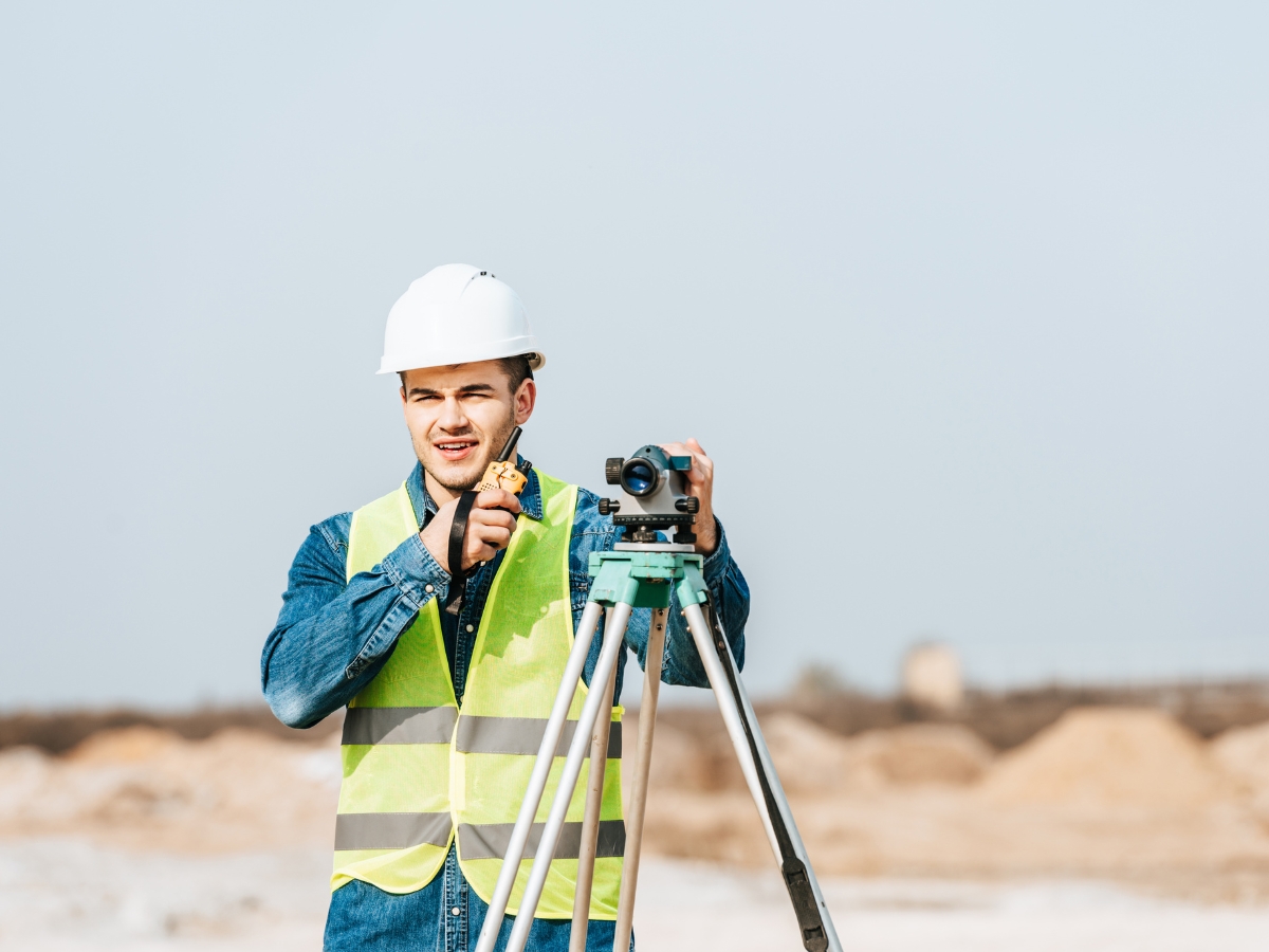 A surveyor in a hard hat and reflective vest uses a surveying instrument on a tripod outdoors.