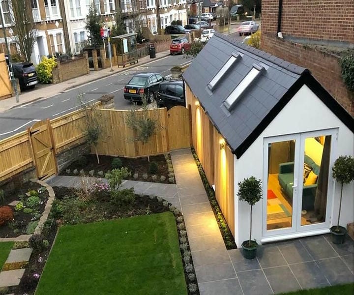 A modern garage conversion with a sleek design, illuminated entrance, surrounded by landscaped gardens and a view of a quiet street.