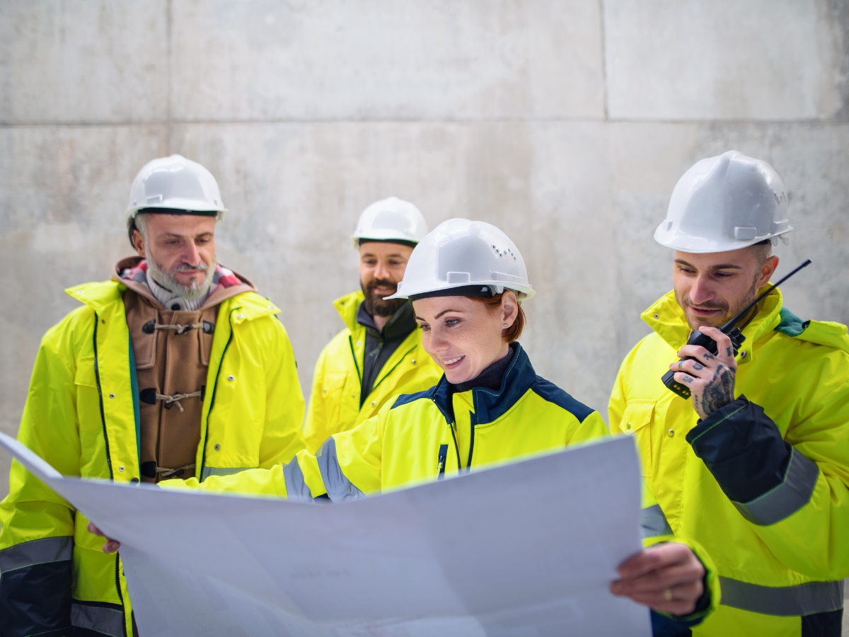 A group engineers in neon yellow safety jackets and white hard hats, reviewing blueprints on a construction site.