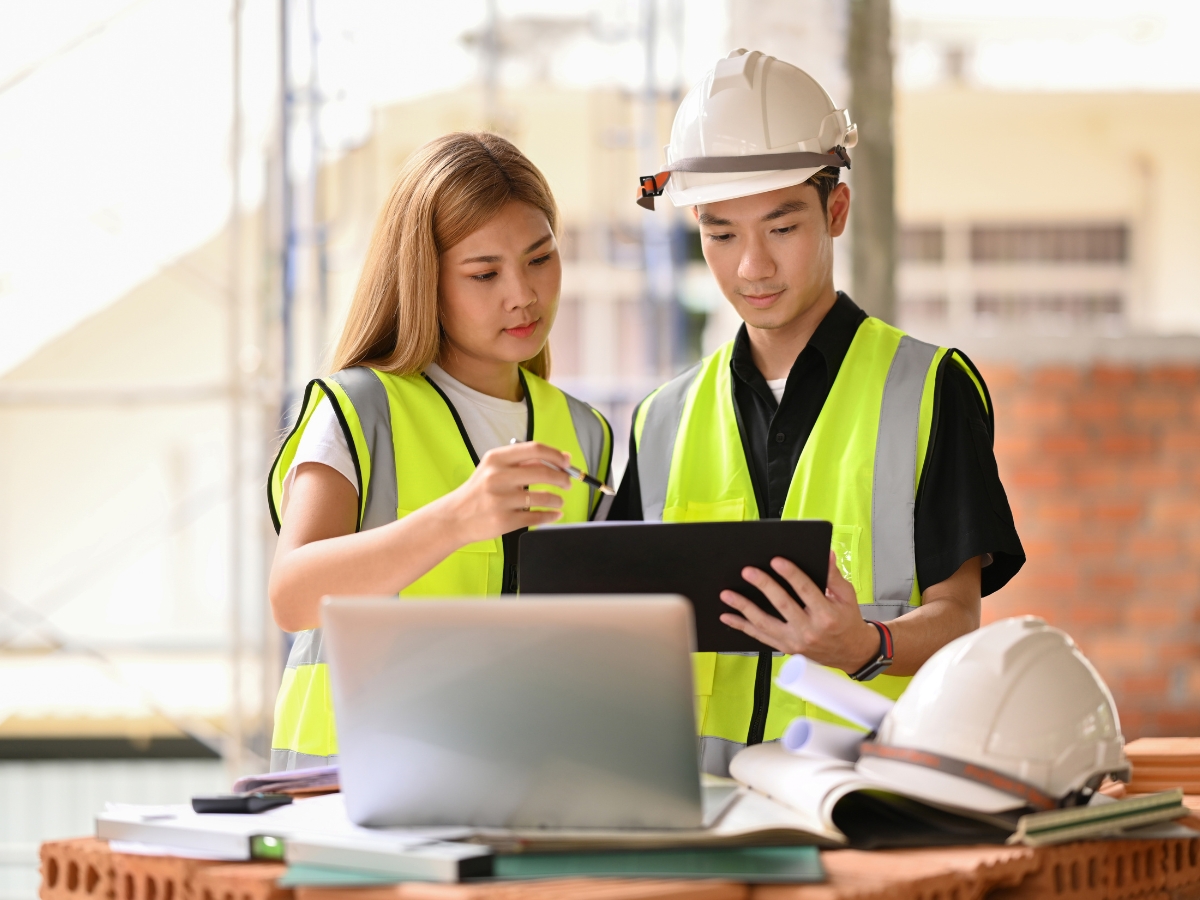 Two architects in bright vests collaborate over a tablet on a worksite, with safety helmets and a laptop on the table.