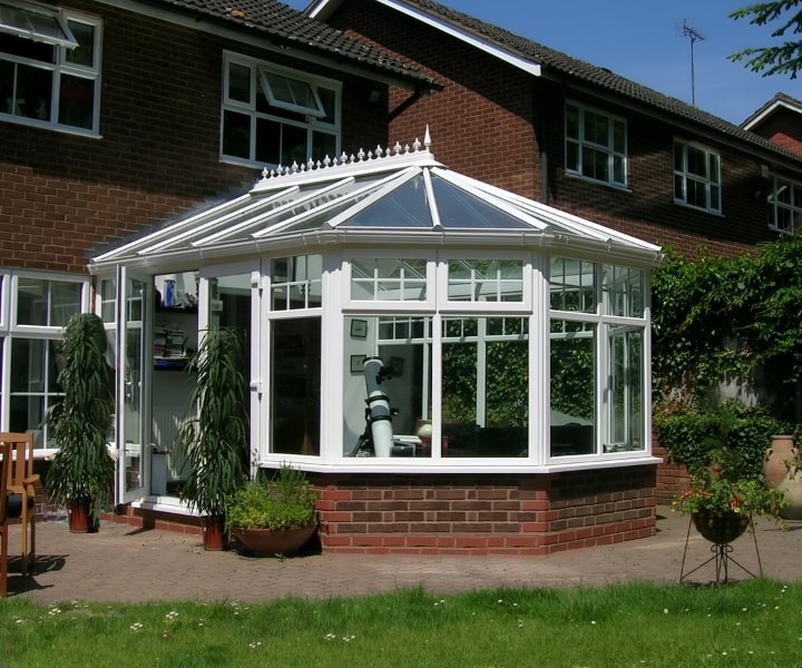 A bright cable conservatory with glass walls and a pointed roof, featuring potted plants and a telescope, attached to a brick house.