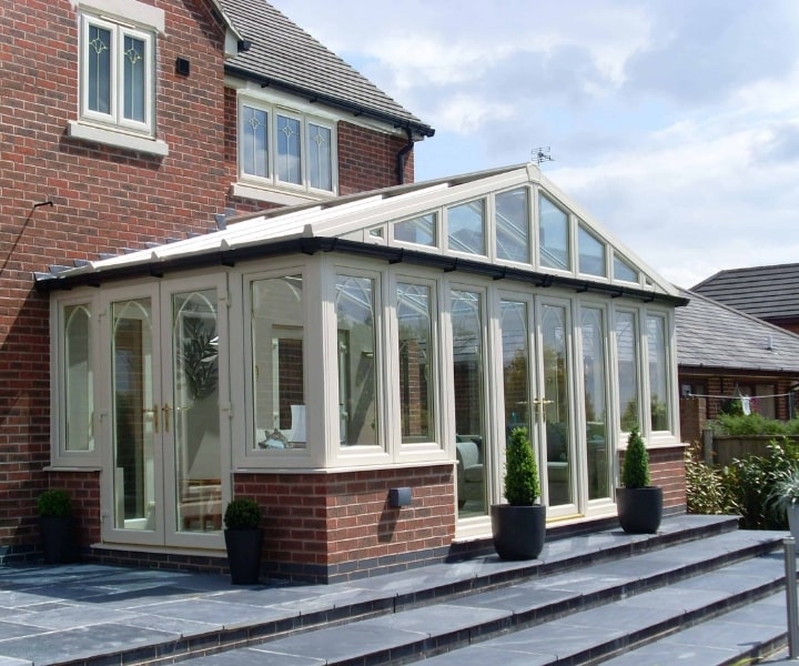 Stylish glass conservatory attached to a brick house, featuring large windows and elegant doors, surrounded by potted plants.