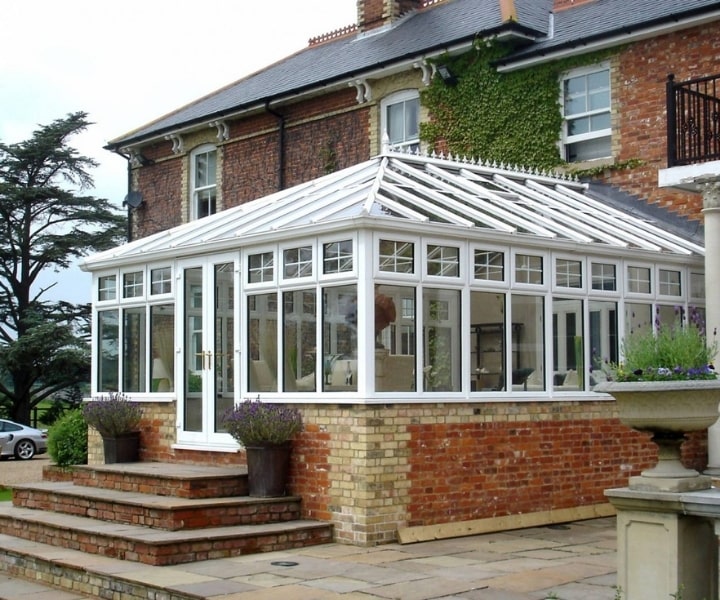 A bright, white-framed conservatory attached to a brick house, featuring large windows and a glass roof, surrounded by greenery.