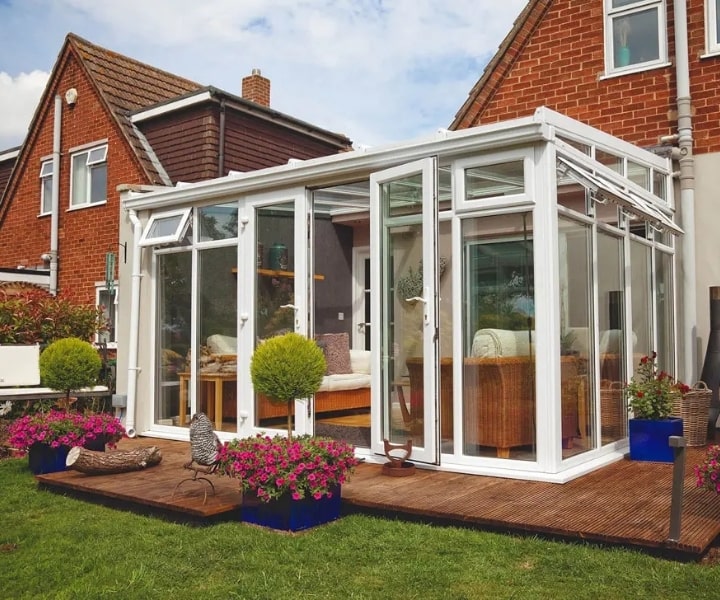 A stunning lean to conservatory with glass walls sits in a garden, surrounded by colorful flowers and green shrubs, beside a cozy home.