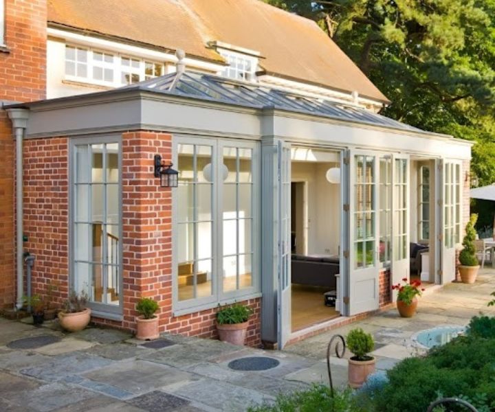 A modern brick conservatory with glass panels, surrounded by potted plants, leading into a light-filled interior.