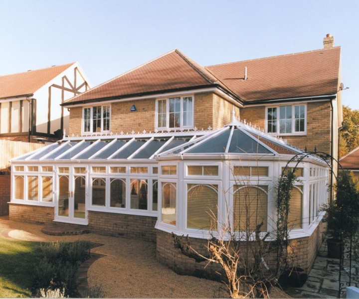 A spacious brick house with a large white P shaped conservatory featuring arched windows and a glass roof, surrounded by a landscaped garden.