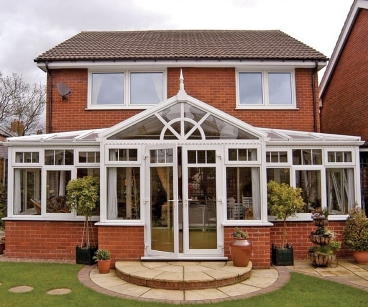 An up to dated red brick house stylised with a spacious white conservatory with large windows, lush plants, and a circular stone patio.