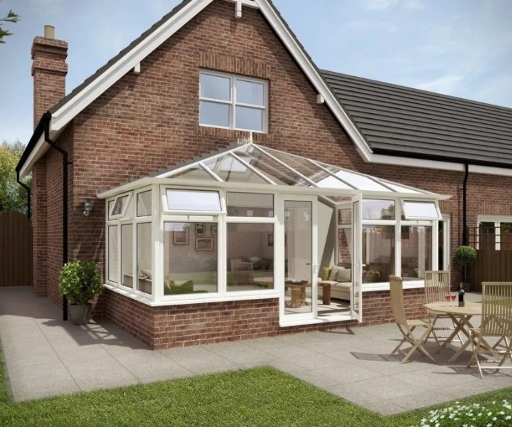 A modern brick house presenting a large glass conservatory and outdoor seating area on a stone patio, surrounded by greenery.