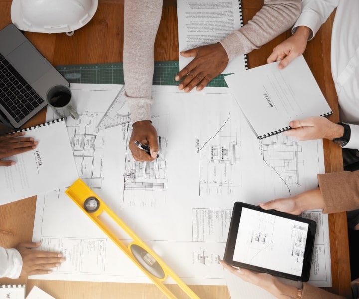 Group of people reviewing architectural blueprints with documents, a laptop, tablet, and measuring tools on the table.