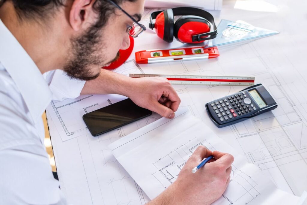 A person working on architectural plans with a calculator, ruler, and measuring tools on a desk