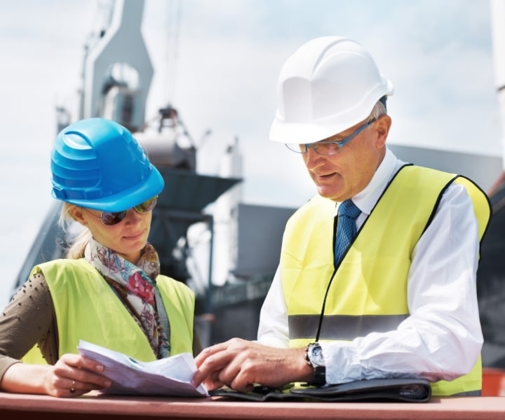 Two engineers in safety vests and hard hats reviewing documents at an industrial site.