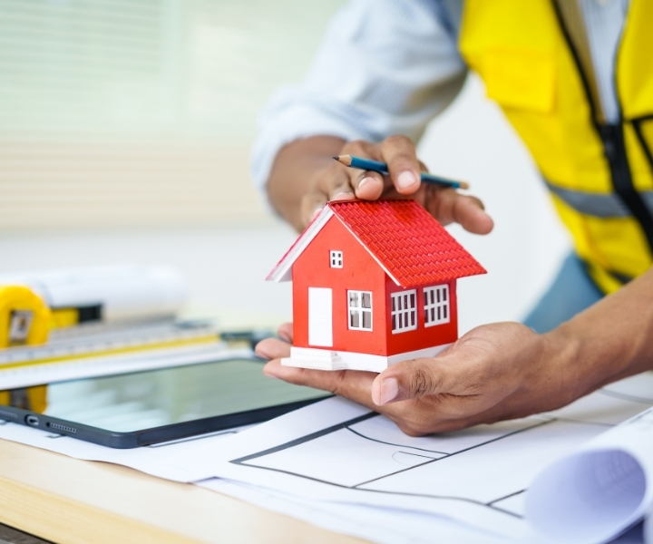 Construction professional holding model house with blueprint and tools.